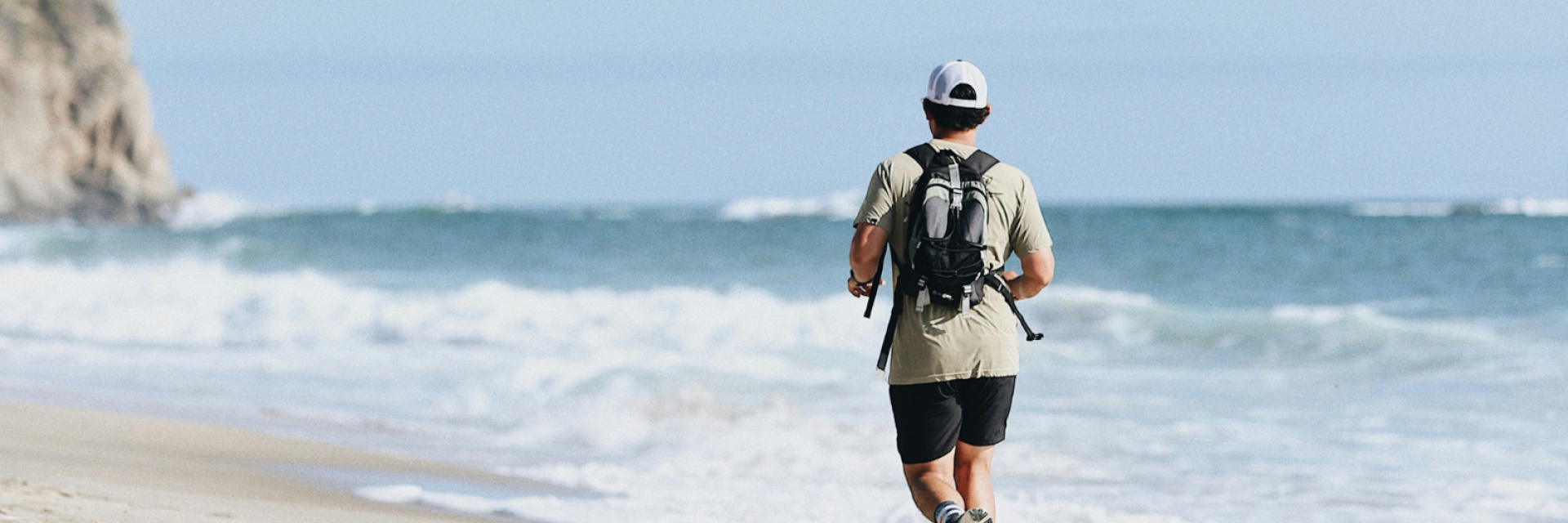Man with a backpack, wearing a cap and jogging along the beach on a sunny day