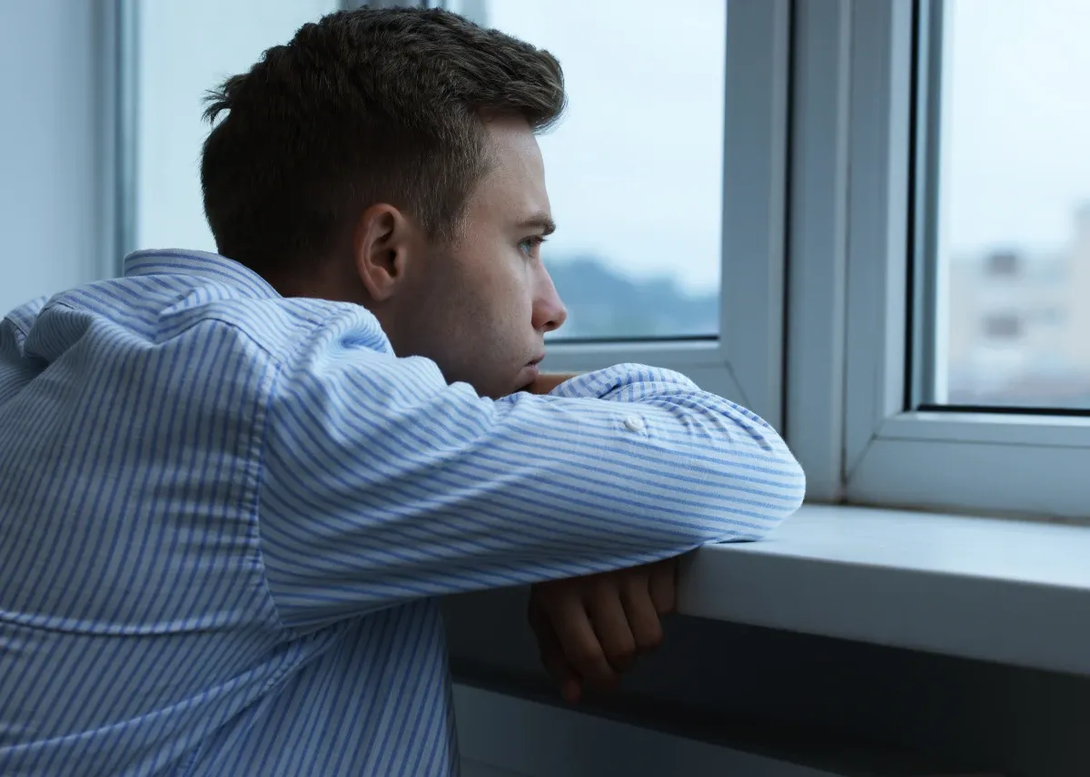 Caucasian male in his 20s, in a blue and white striped shirt, standing with his elbows on a window sill looking out the window