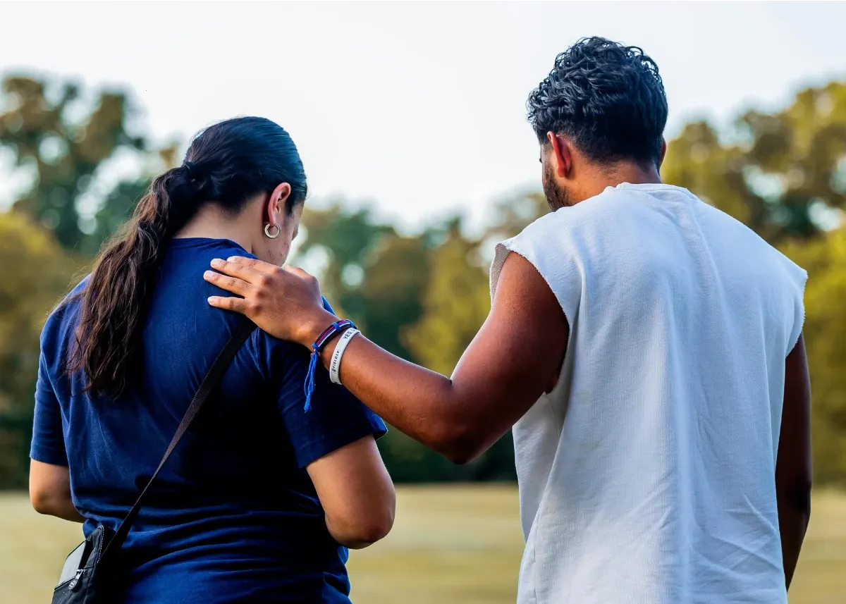 Dark skinned adult man and woman walking and talking in a park setting – the male has his hand resting on the woman's right shoulder in a gesture of support