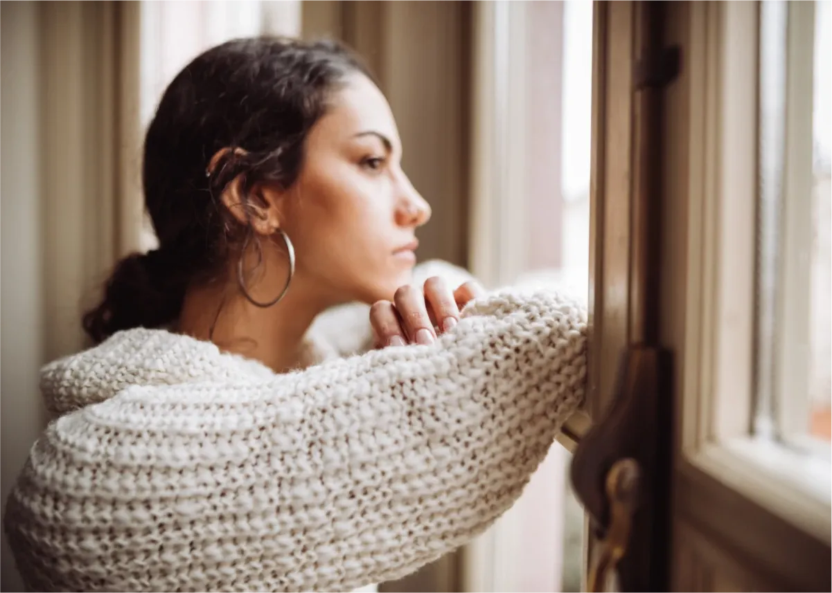 Dark skinned woman with brown hair and brown eyes, wearing a cream chunky woolen jumper and silver hoop earrings, staring out a window and thinking