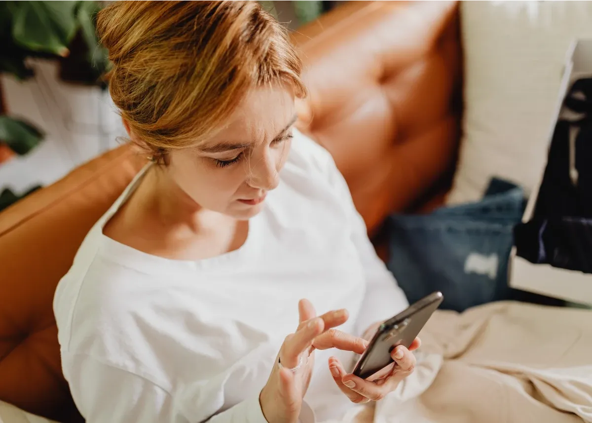 Caucasian woman in her late 30's, with copper hair and a white long sleeved shirt, sitting on a tan couch with a blanket on her lap, navigating SANE Forums on her smartphone.