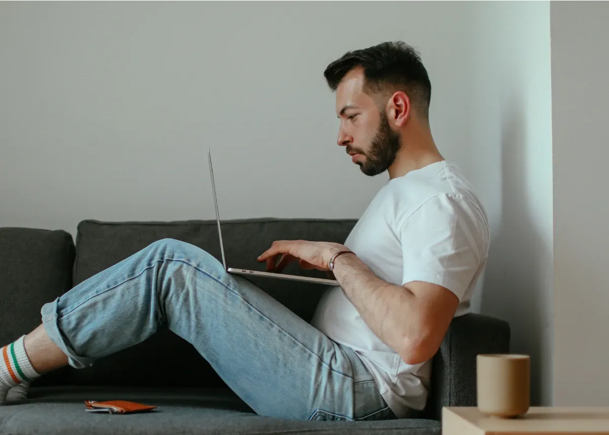 European male in his 30s, in white tshirt, jeans and socks, sitting on a couch, creating a safety plan on his laptop